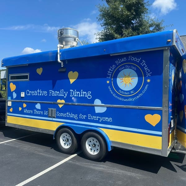 The Little Blue Food Truck, painted blue and yellow with hearts, parked on asphalt.