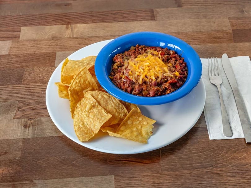 A blue bowl of chili topped with shredded cheese, served with tortilla chips on a white plate.
