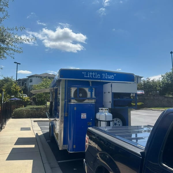 A blue food trailer labeled "Little Blue" is parked next to a black pickup truck on a sunny day.