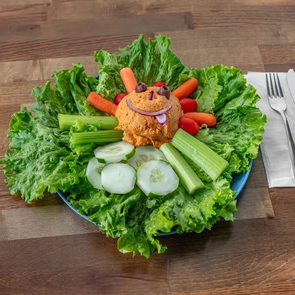 A playful hummus platter shaped like a face with vegetable dippers on a bed of lettuce.